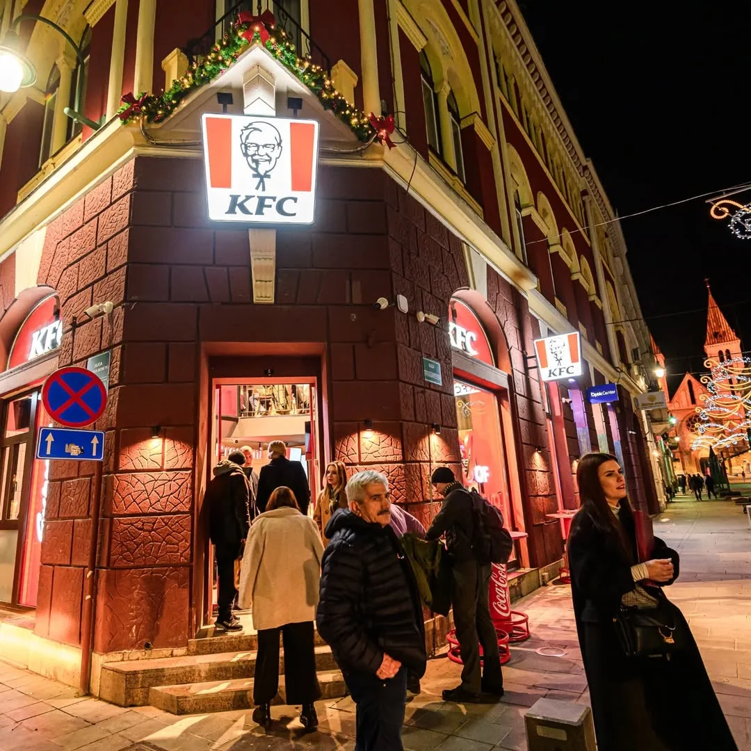 Noćni prikaz ulaza u KFC restoran u ulici Strossmayerova sa osvijetljenim brendom i posjetiocima ispred objekta
